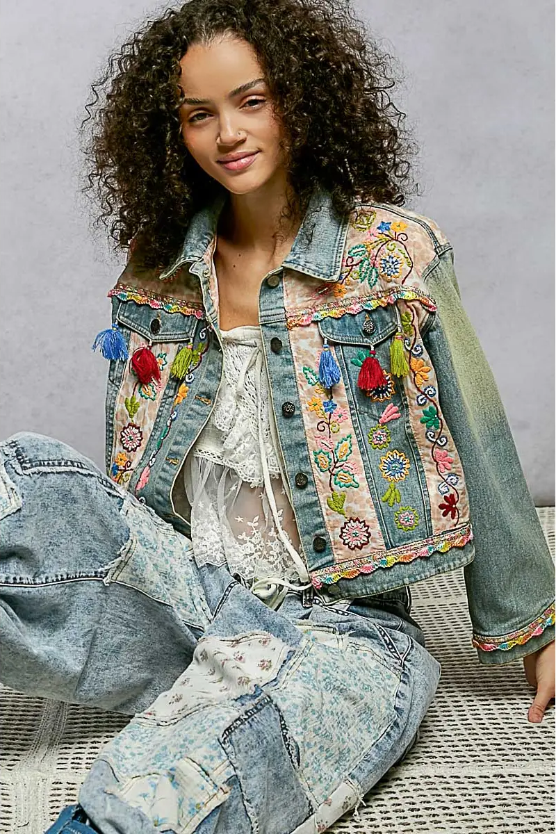 Woman wearing a colorful embroidered denim jacket sitting on a chair.